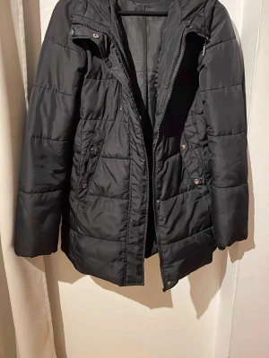 Svart pufferjacka från Vero Moda - Säljer en svart pufferjacka från Vero Moda med huva och dragkedja framtill. Jackan har quiltad design, tryckknappar på fickorna och en skön, lite längre passform som håller dig varm under kyliga dagar.