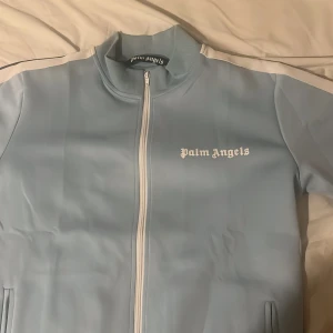 Ljusblå Palm Angels zip hoodie - Säljer en ljusblå zip hoodie från Palm Angels med vita detaljer längs ärmarna och vit dragkedja. Tröjan har hög krage, ribbade muddar och två fickor framtill. Ikonisk Palm Angels-logga på bröstet. Perfekt för en chill och sportig look.