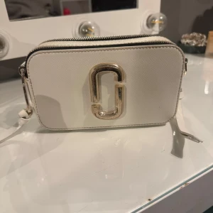 Vit väska från Marc Jacobs - Säljer en stilren vit axelväska från Marc Jacobs med silvriga detaljer som har blivit lite utslitna. Väskan har en avtagbar och justerbar axelrem som har lite små fläckar. Skriv för fler bilder🙌