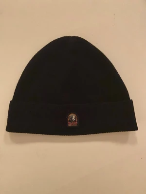 Svart mössa från Parajumpers - Säljer en svart mössa från Parajumpers med uppvikt kant och klassisk logga framtill. Enkel och stilren design som passar till det mesta. Perfekt för kalla dagar. Priset är inte hugget i sten. Skriv vid frågor!