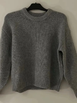 Grå stickad tröja med rund hals - Mysig grå stickad tröja med rund halsringning och ribbade muddar vid ärmslut och nederkant. Tröjan har en loose passform och är perfekt för kyligare dagar. Enkel och stilren design som passar till det mesta. Har små små rosa fillar vid halsringningen kom privat för bilder osv 