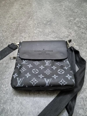 Svart axelrems väska - Snygg svart axelväska med klassiskt monogrammönster i grått. Väskan har justerbar axelrem, magnetlås och dragkedja. Tillverkad i skinn med detaljer i metall. Perfekt för dig som vill ha en stilren och ikonisk accessoar.