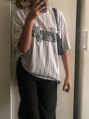 Vit randig oversized t-shirt - Säljer en vit oversized t-shirt med svarta tunna ränder och trycket 'New York' i svart på bröstet. T-shirten har korta ärmar och en klassisk rund halsringning. Perfekt för en chill och sportig look.