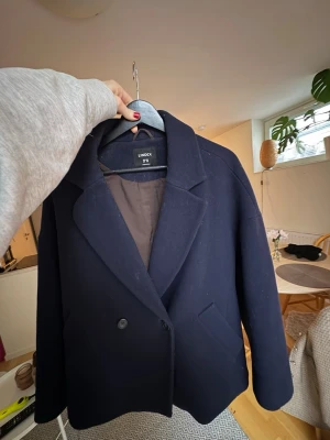 Mörkblå ullkappa från Lindex - Säljer en stilren mörkblå kappa från Lindex i storlek S. Klassisk modell med dubbelknäppning, bred krage och två fickor framtill. Kappan är fodrad och har en rak, något oversized passform som passar perfekt till höst och vinter.