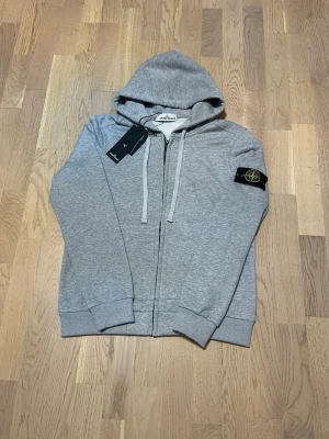 Grå Zip Hoodie - Stone Island - Stilren och ikonisk Stone Island zip-hoodie i klassisk grå färg. Ett premiumplagg med hög kvalitet och tidlös design, utrustad med den välkända avtagbara Stone Island-kompassen på ärmen.  • Märke: Stone Island • Typ: Zip-hoodie • Färg: grå • Passform: S-M • Skick: Mycket fint skick • Avtagbar ikonisk Stone Island-badge på ärmen • Mjuk, tung och högkvalitativ kvalitet  En riktig garderobsfavorit som håller både stil och värde över tid.  🔹 Hög efterfrågan 🔹 Begränsat utbud 🔹 Först till kvarn