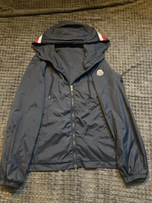 Blå Moncler windbreaker  - Säljer en mörkblå vindjacka från Moncler med huva och dragkedja framtill. Jackan har Moncler-logga på armen och snygga detaljer med röd och vit rand på huvan. Tillverkad i lätt polyester, perfekt för vår och höst. Klassisk passform och elastiska muddar vid ärmslut.