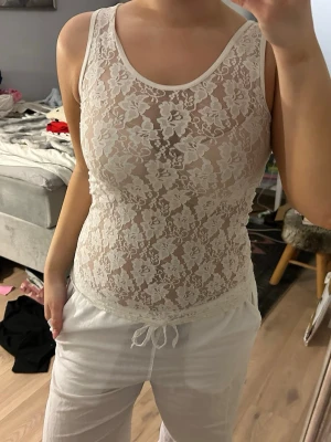 Vit blommig spetslinnetopp - Säljer en vit, ärmlös linne-topp i genomskinlig spets med blommigt mönster. Toppen har rund halsringning och är figurnära. Perfekt för lager på lager eller som statement-plagg.