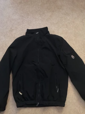 Svart Moncler bomberjacka M - Den är ny och har använt den 5 gånger Svart bomberjacka från Moncler med klassisk ståkrage, två fickor med dragkedja och en ficka på ärmen med Moncler-logga. Jackan är tillverkad i polyester och har en stilren, loose passform. Perfekt för dig som vill ha en clean look med premiumkänsla.