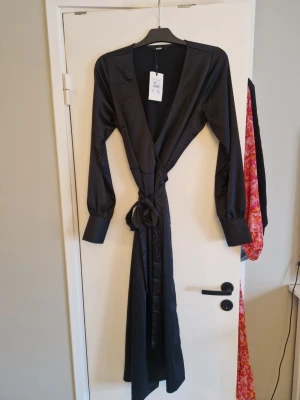 Svart omlottklänning från Bik Bok - Snygg svart långklänning från Bik Bok i glansigt satintyg. Klänningen har omlottmodell med knyt i midjan, djup v-ringning och långa ärmar med breda manschetter. Perfekt för dig som vill ha en elegant och stilren look.