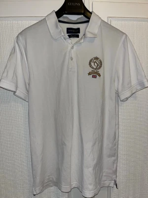 Vit pikétröja från Henri Lloyd - Klassisk vit pikétröja från Henri Lloyd med broderad logga och jubileumsmärke på bröstet. Tröjan har krage, korta ärmar och knappar framtill. Tillverkad i bomull för en skön och luftig känsla.