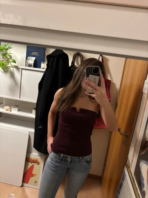 Vinröd bandeau tubtopp - Säljer en vinröd bandeau tubtopp med en snygg liten v-formad detalj framtill. Toppen är axelbandslös och har en tight passform som sitter på plats. Materialet känns stretchigt och mjukt, perfekt att styla med jeans eller kjol.