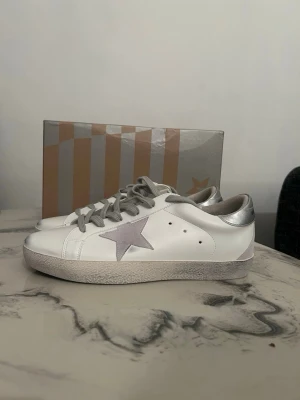 Golden Goose vita sneakers med silverdetaljer - Golden Goose sneakers i vitt skinn med silverfärgad häl och grå stjärndetalj på sidan. Skorna har grå snörning, platt sula och svart yttersula. Klassisk låg modell med rund tå och diskret branding på hälen.