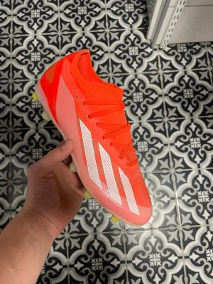 Adidas orange fotbollsskor strl 38⅔ - Säljer ett par grymt snygga orangea Adidas fotbollsskor med vita ränder och texten CRAZYFAST på sidan. Skorna har snörning, platt sula med gula dobbar och är tillverkade i syntetmaterial. Perfekta för dig som vill sticka ut på planen!