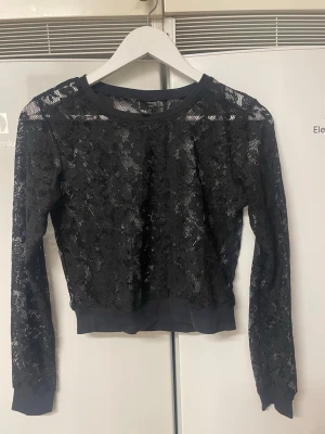 Svart topp i svart spets - Säljer en svart croppad blus i genombruten spets med blommönster. Toppen har långa ärmar, rund halsringning och breda muddar vid ärmslut och nederkant. Perfekt för dig som vill ha en edgy och trendig look.