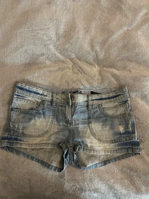 Jeans shorts  -  Snygga jeansshorts för flicka i ljusblå tvätt med trendig, sliten look. Shortsen har en bekväm passform och är perfekta för både vardag och sommaraktiviteter.  Framsida: Klassisk jeansdesign med två framfickor och myntficka. Knapp- och dragkedjestängning samt bälteshällor. Dekorativa slitningar ger en modern och cool stil.  Baksida: Två bakfickor med lock och knapp som ger en snygg detalj och extra karaktär. Lätt blekt finish för ett trendigt utseende.  Plagget är i fint begagnat skick och pass