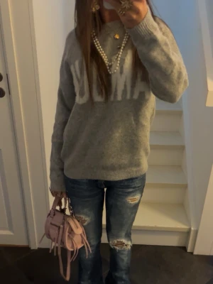 Grå stickad tröja med vit text - Mysig grå stickad tröja med stor vit text framtill. Tröjan har rund halsringning och långa ärmar. Perfekt för dig som gillar en enkel men ändå statement look. Passar till jeans eller mjukisbyxor för en chill vibe.