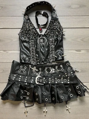 Gotisk topp och minikjol - Svart set med en halterneck- topp dekorerad med nitar och en matchande minikjol med detaljer som ringar, nitar och korsberlocker.