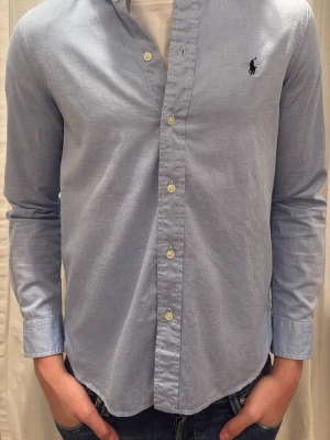 Ljusblå skjorta från Ralph Lauren - Klassisk ljusblå skjorta från Ralph Lauren med lång ärm och knappar framtill. Skjortan har den ikoniska broderade loggan på bröstet och är tillverkad i mjuk bomull. Perfekt passform som sitter snyggt till jeans eller chinos. Storlek 170 men modellen är 185 cm