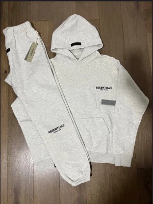 FOG Essentials ”Light Oatmeal” set SS23 - Ljusgrå hoodie från Essentials Fear of God med svart logga på bröstet och ryggen. Klassisk huva, känguruficka och ribbade muddar. Tillverkad i mjuk bomullsmix för maximal komfort och stilren look. Passerar StockX och GOAT‼️
