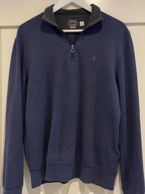 Marinblå tröja från Polo Ralph Lauren - Snygg marinblå långärmad tröja från Polo Ralph Lauren med halv dragkedja och diskret logga på bröstet. Mjuk bomullsblandning och klassisk passform, perfekt till höst och vår. Enkel och stilren design som funkar till det mesta.