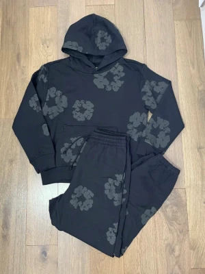 Denim Tears The Cotton Wreath Triple Black Tracksuit - Säljer ett svart set med hoodie och byxor, dekorerat med stora blommiga tryck i grått. Denna Denim Tears är StockX och GOAT passerande och verifierade. Alla sizes tillgängliga.
