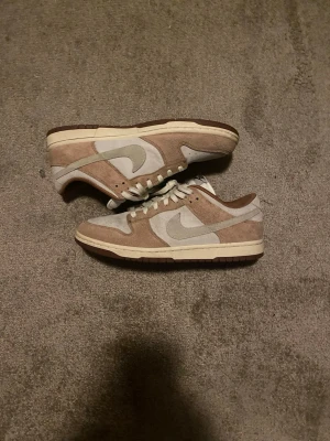 Nike Dunk Low Medium Curry - EU 45 - Nike Dunk Low sneakers i beige och ljusgrå mocka med vita detaljer och bruna sulor. Klassisk Nike Swoosh på sidan och broderad Nike-logga bak. Skorna har snörning och låg profil, perfekt för dig som gillar stilrena och neutrala färger.