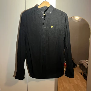 Svart manchesterskjorta Lyle & Scott  - Svart skjorta i mjuk manchester från Lyle & Scott, med klassisk krage och knappstängning. På bröstet finns en ficka med den ikoniska gula fågeln broderad. Skjortan har lång ärm och en stilren, normal passform som funkar till många olika looks.