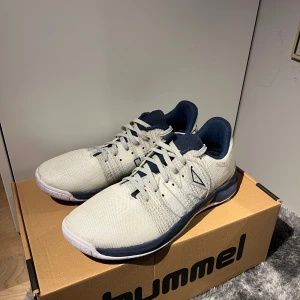 Vita och marinblå träningsskor ifrån kempa  - Snygga träningsskor från Kempa i vitt och marinblått med sportig design. Ovandelen är i slitstark syntet med ventilerande mesh och snörning framtill. Sulan är vit med blå detaljer och har bra grepp, perfekt för inomhussport. Skorna har rund tå och bekväm stoppning runt ankeln. Använda under en träning och säljer för de inte passar foten. HELT NYA 