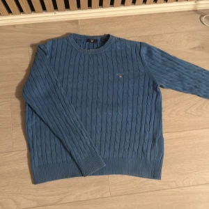 Blå kabelstickad tröja från GANT - Snygg blå kabelstickad tröja från GANT med rund hals och diskret logga broderad på bröstet. Tröjan har ribbade muddar vid ärmslut och nederkant, och är långärmad. Perfekt för dig som gillar klassisk stil med en modern touch.