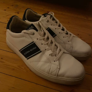 Vita sneakers från Björn Borg - Snygga vita sneakers från Björn Borg med marinblå detaljer och logga på sidan. Skorna har klassisk snörning, rund tå och är tillverkade i ett slitstarkt syntetmaterial med struktur. Perfekta för dig som gillar en clean och sportig stil.
