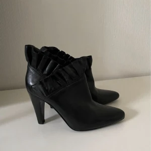 Svarta stövletter med volangdetalj skinn/läder - Snygga svarta stövletter i skinn med hög klack. Skorna har en unik volangdetalj runt skaftet som ger en cool och trendig look. Perfekta för dig som vill sticka ut med dina skor och gillar en elegant men edgy stil.