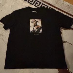 Svart Tupac t-shirt med tryck - Svart t-shirt med ett ikoniskt Tupac-tryck på bröstet. Klassisk rund hals och korta ärmar. Perfekt för dig som vill ha en streetstyle vibe och gillar hiphop. Materialet känns som bomull och passformen är normal.