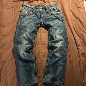 Rock Revival jeans med snygga detaljer - Snygga blå jeans från Rock Revival med feta detaljer. Jeansen har raka ben, kontrastsömmar och detaljerade knappar. Tillverkade i 100% bomull. W31 L30