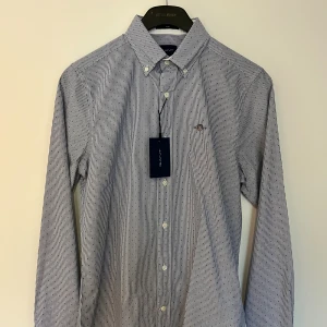 Randig blå skjorta från Gant S - Snygg slim fit skjorta från Gant i storlek S, med blå och vita ränder samt små prickar. Klassisk button-down krage, broderad Gant-logga på bröstet och vita knappar. Tillverkad i bomull och perfekt för en clean och stilren look.
