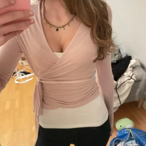 Ljusrosa omlottopp med knyt - Säljer en ljusrosa omlottopp med långa ärmar och snygg v-ringning i storlek xs. Toppen har ett knyt i midjan som ger en smickrande siluett och är tillverkad i ett mjukt, stretchigt material. Perfekt att styla med jeans eller kjol för en trendig look.