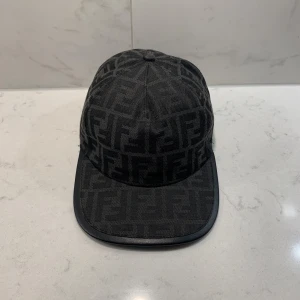 Svart keps från Fendi med logomönster - Snygg svart keps från Fendi med ikoniskt FF-logomönster över hela kepsen. Justerbar baktill med Fendi-detalj. Tillverkad i polyester och bomull för en bekväm och stilren look. Perfekt accessoar för att lyfta din outfit. den är ganska ny och sitter perfekt 