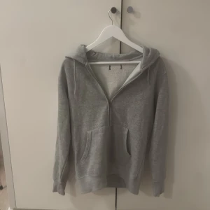 Grå hoodie med dragkedja och huva - En basic grå hoodie med dragkedja framtill och huva. Tröjan har två fickor på magen och långa ärmar. Perfekt för chill dagar eller när du vill ha något bekvämt och enkelt. Gjord i mjukt material med ribbade muddar vid ärmslut och nederkant.
