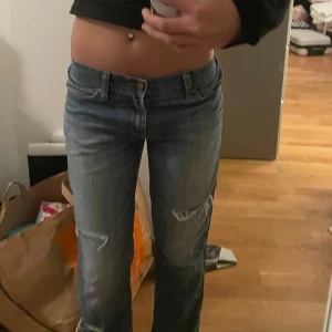 Blå lågmidjade jeans med slitningar - Säljer ett par blå bootcut jeans med låg midja och slitna detaljer på benen. Jeansen har klassisk femficksdesign och är tillverkade i jeansmaterial. Perfekt för dig som gillar en avslappnad och cool stil. Innerbenslängd 79cm & midjemått 39