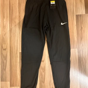 Svarta träningsbyxor från Nike - Svarta träningsbyxor från Nike med klassisk vit swoosh-logga på benet. Byxorna har elastisk midja och är tillverkade i ett lätt och smidigt syntetmaterial som passar perfekt för sport och träning. Avsmalnande ben och enkel, stilren design.