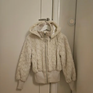 Vit teddyjacka med huva från Kaos - Mysig teddyjacka i vit färg från Kaos by Auluna, storlek 36. Jackan har huva, ribbade muddar och midja, samt dragkedja framtill. Utsidan är fluffig och insidan har ett lent, satinliknande foder. Perfekt för kalla dagar och riktigt snygg till jeans.