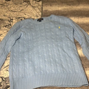 Ljusblå kabelstickad tröja Polo Ralph Lauren - Kabelstickad ljusblå tröja från Polo Ralph Lauren med rund halsringning och klassisk gul broderad logga på bröstet. Tröjan har långa ärmar och ribbade muddar vid ärmslut och nederkant. Perfekt för en stilren och chill look.