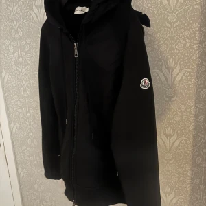 Moncler Cardigan  - Svart cardigan från Moncler med dragkedja framtill, huva och klassisk logga på ärmen.  Passar bra på mig och är runt 176, de finns en kod som bevisar att den är äkta, skickar den bilden när intresse finns!