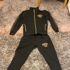 Svart tracksuit med guldtiger - Snygg svart tracksuit med guldfärgad tigertryck på både jacka och byxa. Jackan har dragkedja framtill och två fickor med guldfärgade detaljer. Ärmarna har dekorativa ränder. Perfekt för dig som gillar sportig och trendig stil.