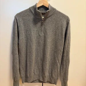 Oscar Jacobson Zip - Klassisk grå stickad tröja från Oscar Jacobson med ribbad krage och half zip-dragkedja. Tillverkad i Italien och har långärmad design. Perfekt för lager-på-lager under kyliga dagar.