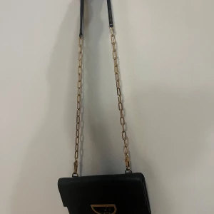 Svart axelväska från Coccinelle - Snygg svart axelväska från Coccinelle i skinn med guldfärgad kedja och metallspänne. Väskan har en stilren, boxig form och elegant struktur på ytan. Perfekt för dig som gillar klassiska detaljer och vill ha en väska med lyxig känsla.