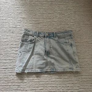 Ljusblå jeanskjol med fickor - Säljer en ljusblå jeanskjol i klassisk modell med fem fickor, bälteshällor och knappstängning framtill. Kjolen är kort och har en rak passform med rå kant nertill för en avslappnad look. Perfekt till sneakers eller boots.