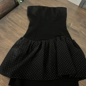 Svart prickig kort klänning från Maje - Supersnygg svart axelbandslös kort klänning från Maje med voluminös kjol i prickigt tyg. Klänningen har en figurnära överdel och en cool volangdetalj i midjan. Perfekt för dig som vill sticka ut med en chic och modern look.