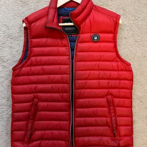 Röd dunväst från Sail Racing - Snygg röd dunväst från Sail Racing med quiltad design och ståkrage. Västen har två fickor med dragkedja och ett Sail Racing-märke på bröstet. Insidan är mörkblå och materialet är lätt och smidigt, perfekt för lager-på-lager. Passar dig som gillar sportig och clean stil.