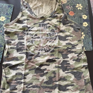 Camouflage linnetopp med tryck stonen island - Säljer en cool ärmlös linnetopp med klassiskt camouflage-mönster i grönt, beige och svart. Toppen har ett stort tryck med kompassmotiv och text framtill. Perfekt för dig som gillar streetstyle och vill sticka ut med militärinspirerad look.