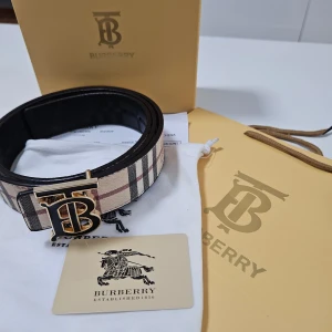 Burberry randigt bälte med guldspänne - Snyggt Burberry-bälte med klassiskt randigt mönster i beige, svart och rött. Bältet har ett exklusivt guldigt 'TB'-spänne och är tillverkat i skinn. Insidan har ett diskret rutigt mönster i svart. Perfekt accessoar för att lyfta din outfit.